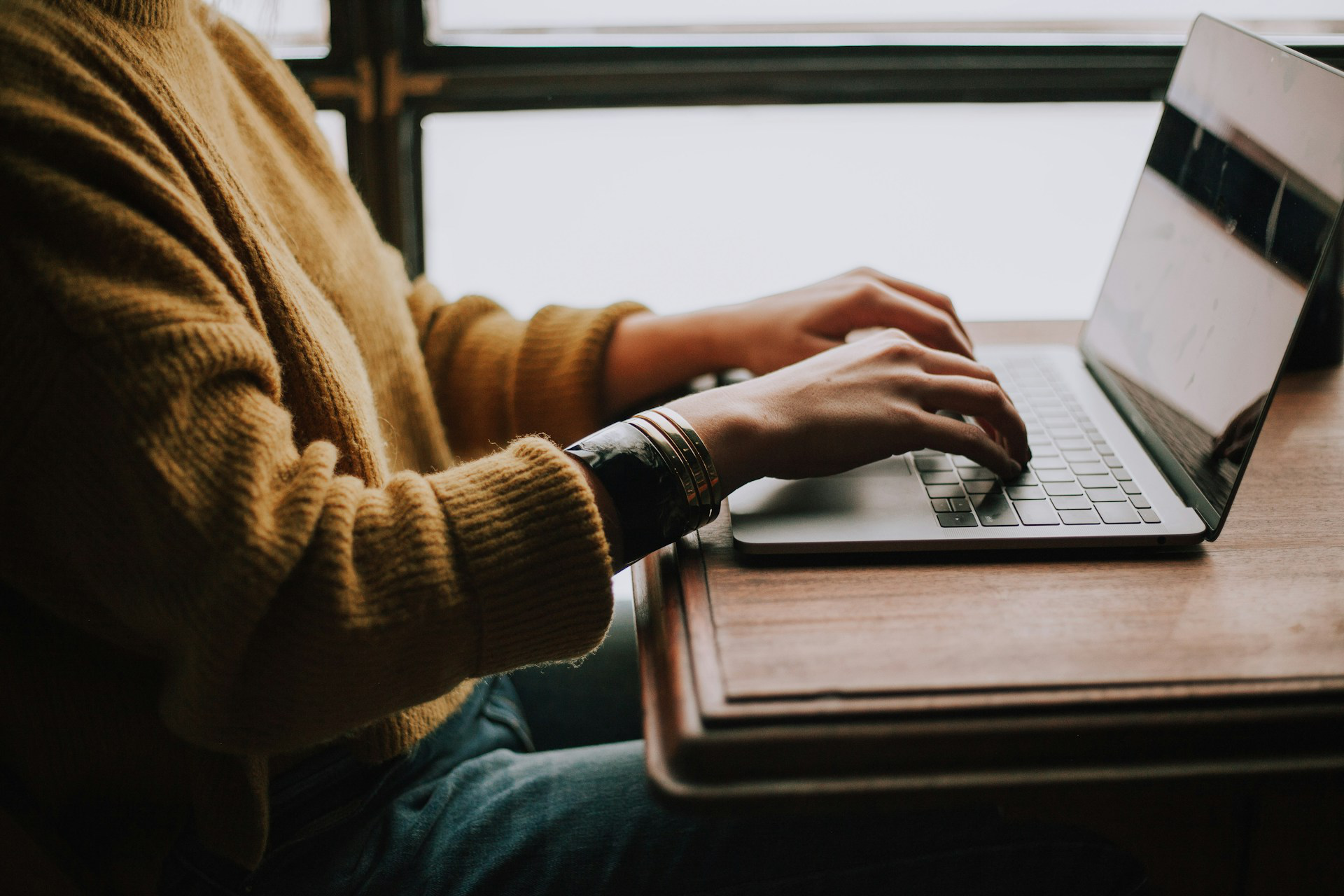 A picture of a person wearing yellowish brown sweater, typing on a mac laptop.